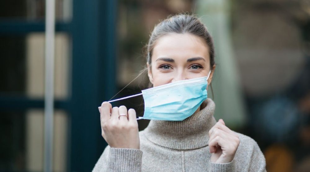 Portrait of a young attractive Caucasian woman standing outdoors and smiling while putting on a protective face mask
